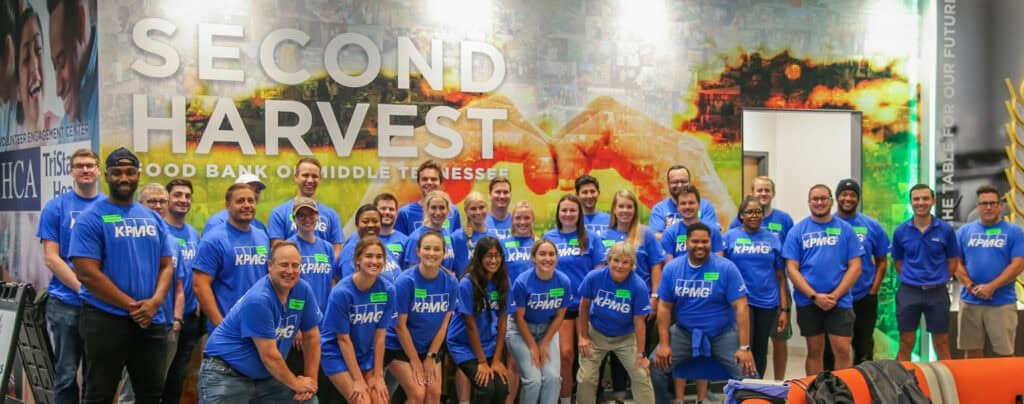 A large group of people in matching blue KPMG shirts pose and smile together in front of a mural wall reading Second Harvest Food Bank of Middle Tennessee.