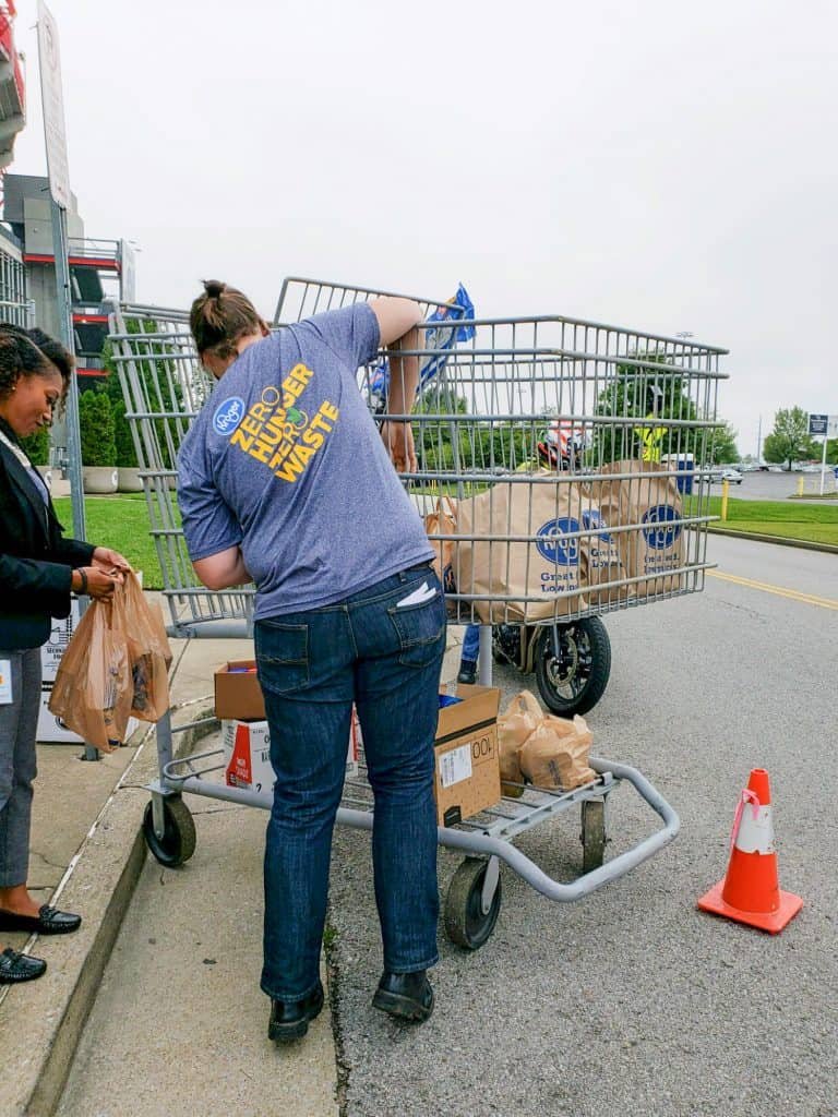 A person in a blue shirt loading groceries into a large metal cart next to a curb, while another person stands nearby holding bags. There are cardboard boxes and paper bags in the cart and an orange traffic cone on the ground.