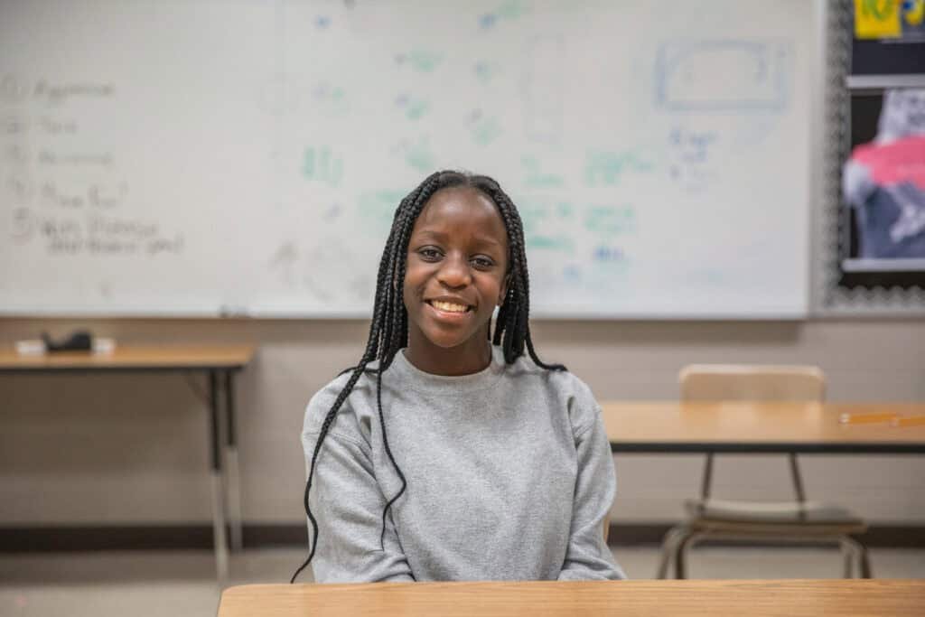 A young woman with long braided hair, wearing a gray sweatshirt, sits and smiles at a desk in a classroom with a whiteboard and empty desks behind her.