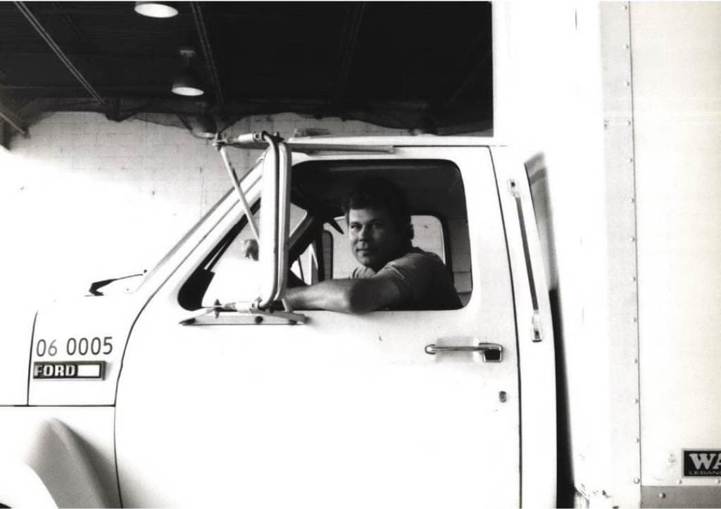 A man sits in the driver’s seat of a white Ford truck, looking out the open window. The truck is parked indoors near a white wall, and the area is dimly lit by overhead lights.
