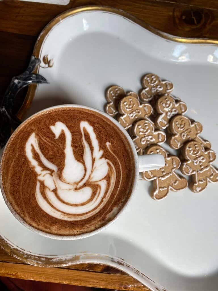 A cup of coffee with swan latte art sits on a tray next to several gingerbread cookies shaped like people, all decorated with white icing.