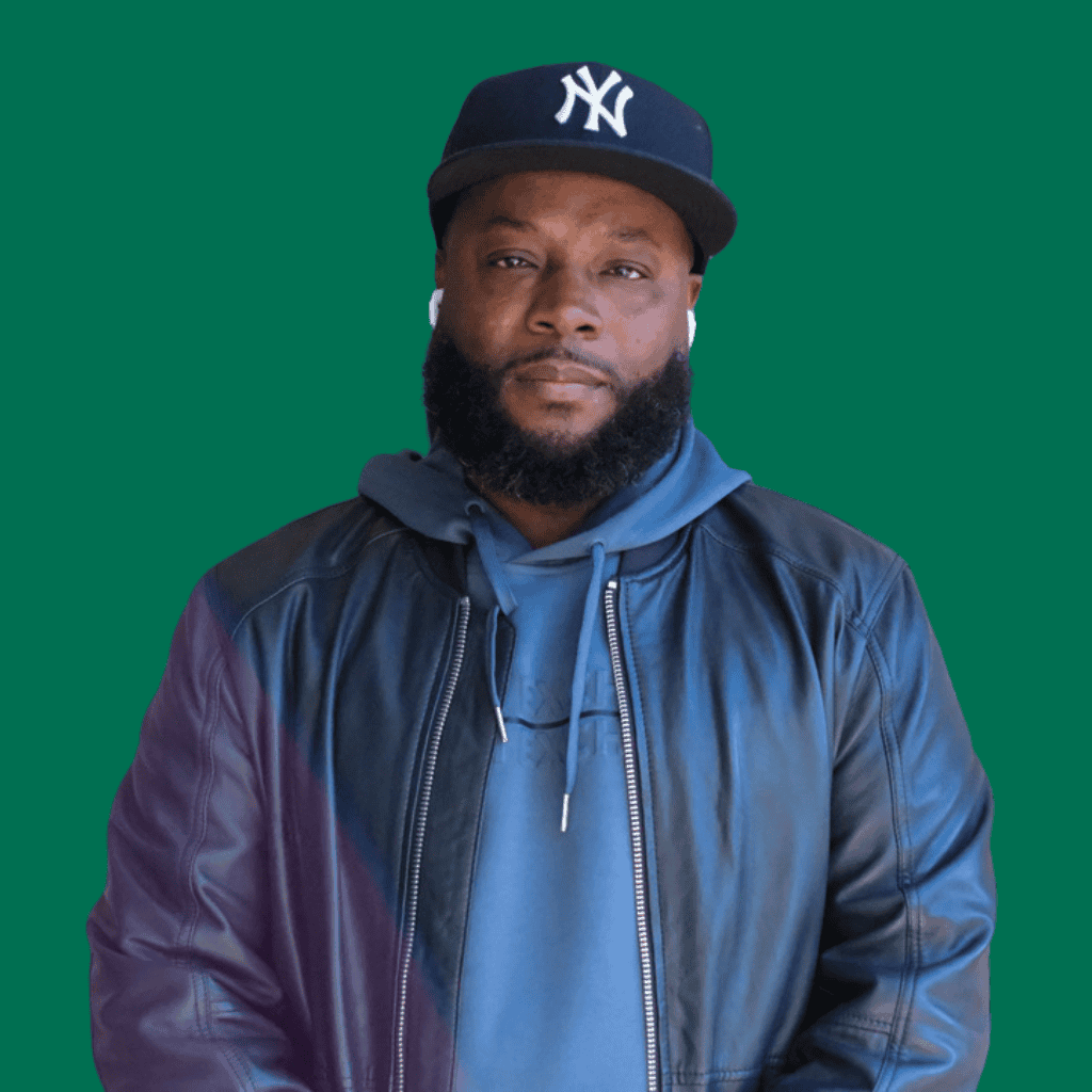 A man with a beard wearing a black New York Yankees cap, wireless earbuds, a blue hoodie, and a black leather jacket, stands against a solid green background.