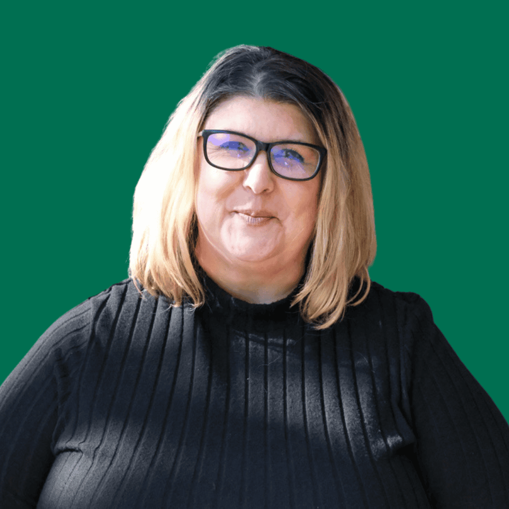 A woman with shoulder-length blonde hair and glasses, wearing a black ribbed top, smiles in front of a solid green background.