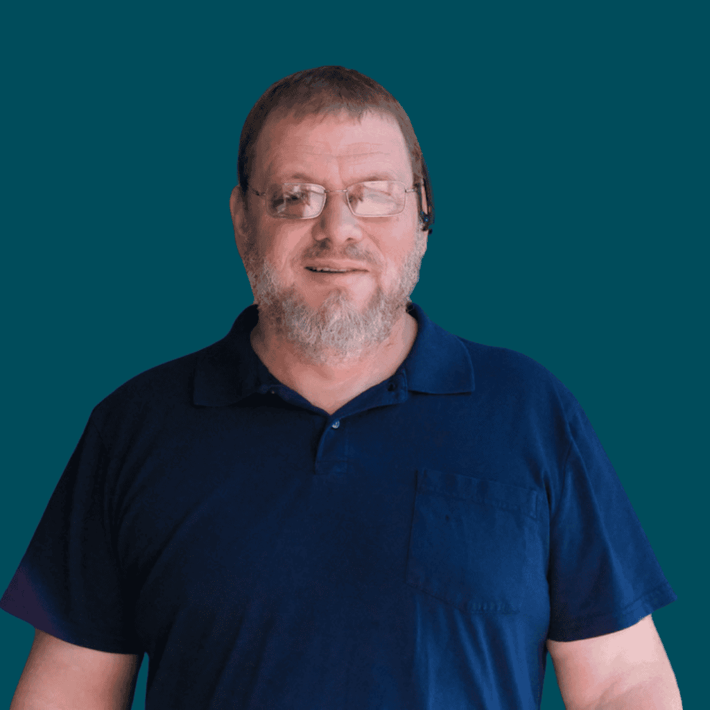 A middle-aged man with short hair, glasses, and a beard is smiling slightly. He is wearing a dark blue polo shirt and standing against a solid teal background.