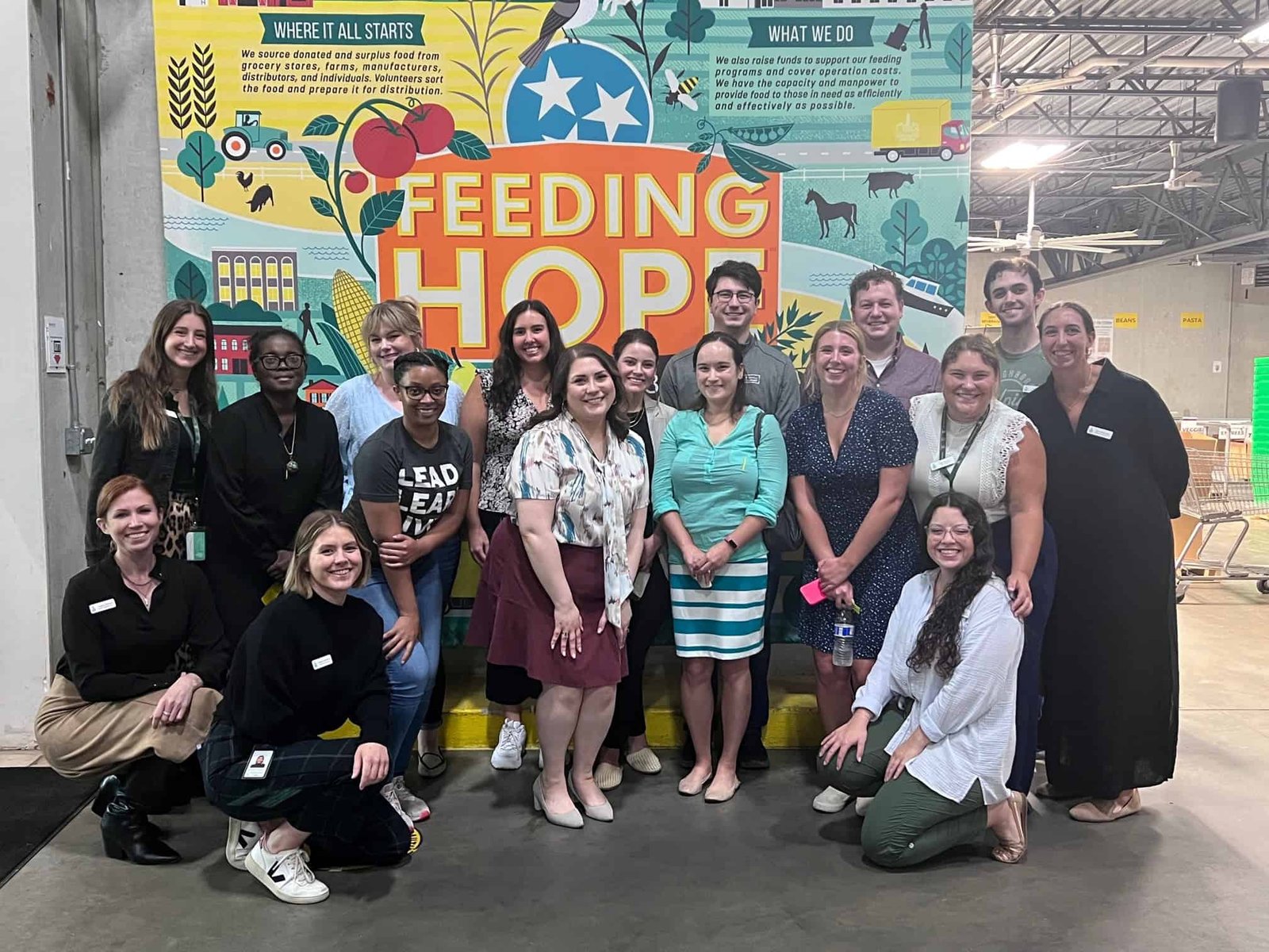 A group of people pose and smile in front of a colorful mural that says Feeding Hope inside an industrial building, suggesting teamwork or volunteering at a food bank or community center.