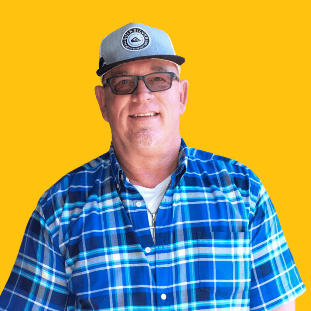 Smiling man wearing glasses, a gray baseball cap, and a blue plaid shirt poses in front of a bright yellow background.