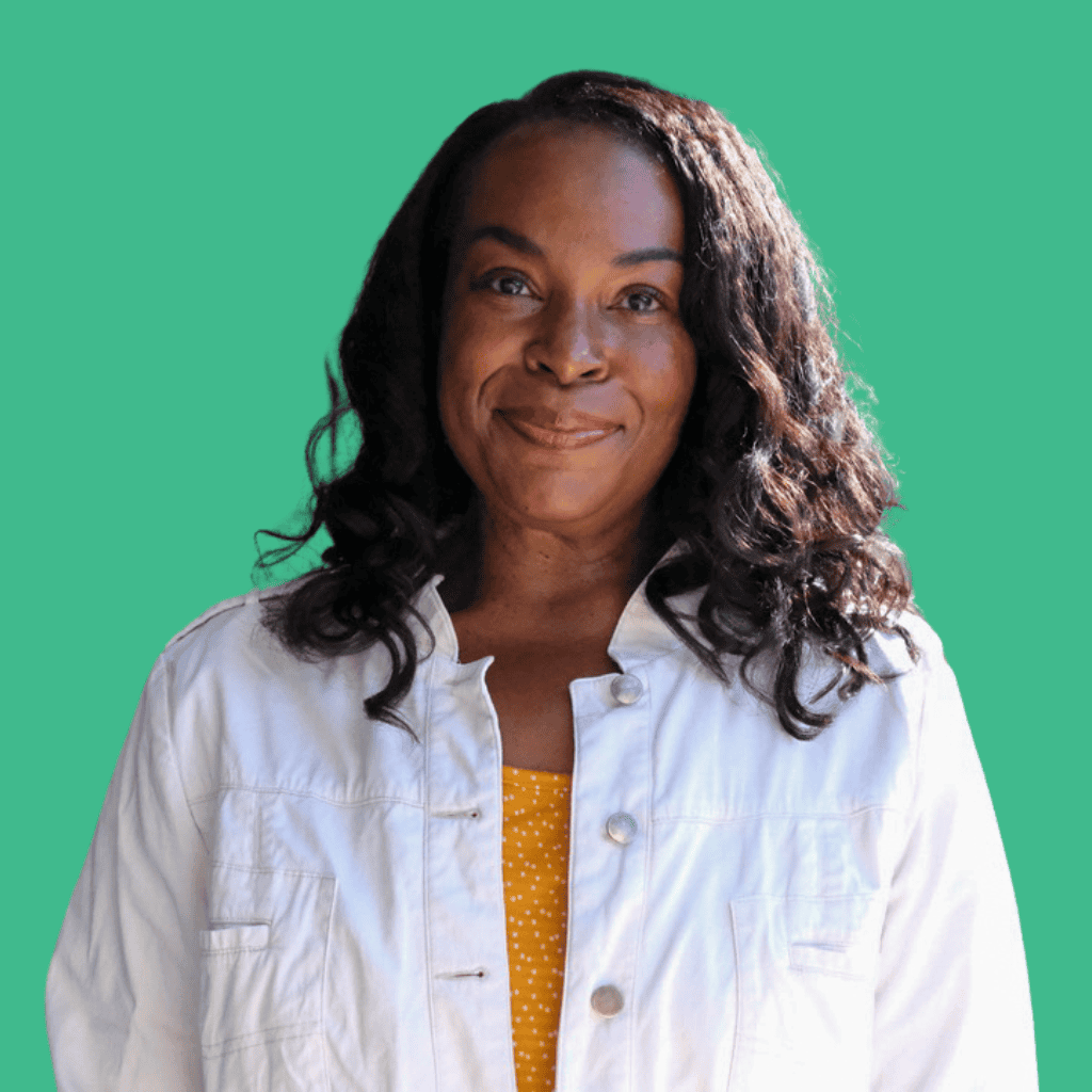 A woman with long curly hair smiles gently while wearing a white jacket over a yellow top with white polka dots, standing in front of a solid teal background.