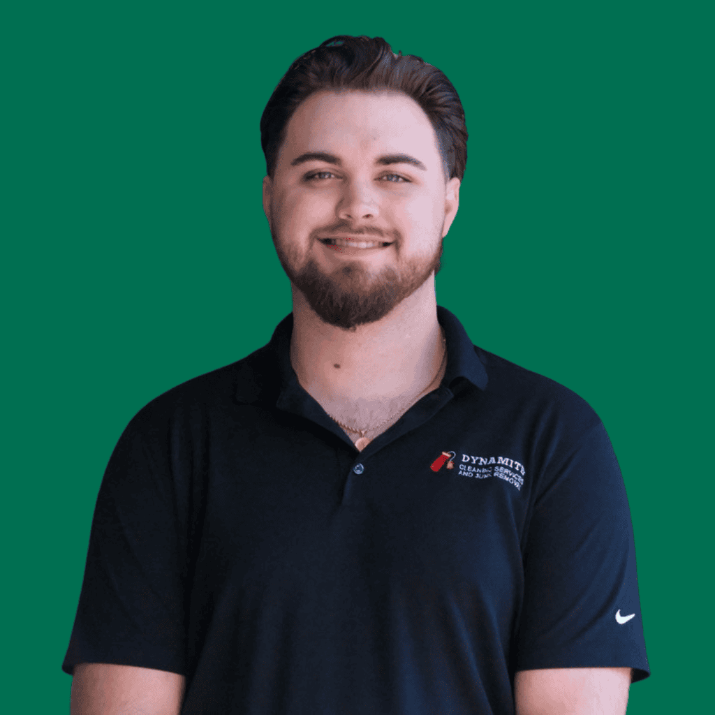 A man with short brown hair and a beard, wearing a black collared shirt with a DYNAMITE logo, stands smiling in front of a solid green background.