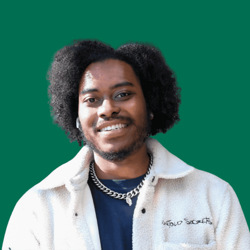 A young man with curly hair and a beard smiles at the camera, wearing a cream fleece jacket, a chain necklace, and a dark shirt, standing in front of a solid green background.
