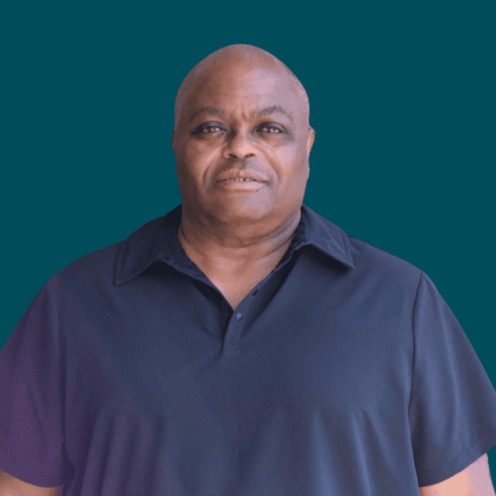 A middle-aged man with short hair wearing a navy blue collared shirt stands in front of a solid teal background, looking at the camera with a neutral expression.