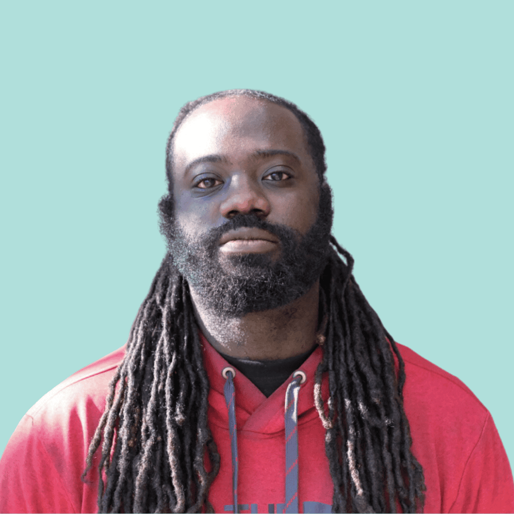 A man with long, dark dreadlocks and a full beard wears a red hoodie and looks straight at the camera against a plain light blue background.