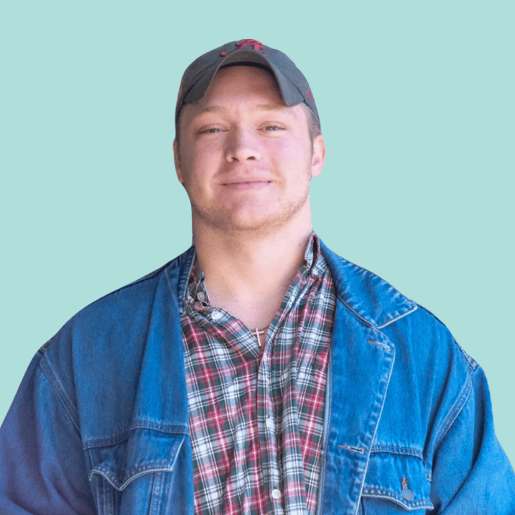 A man wearing a green baseball cap, a red and green plaid shirt, and a blue denim jacket stands smiling in front of a light blue background.