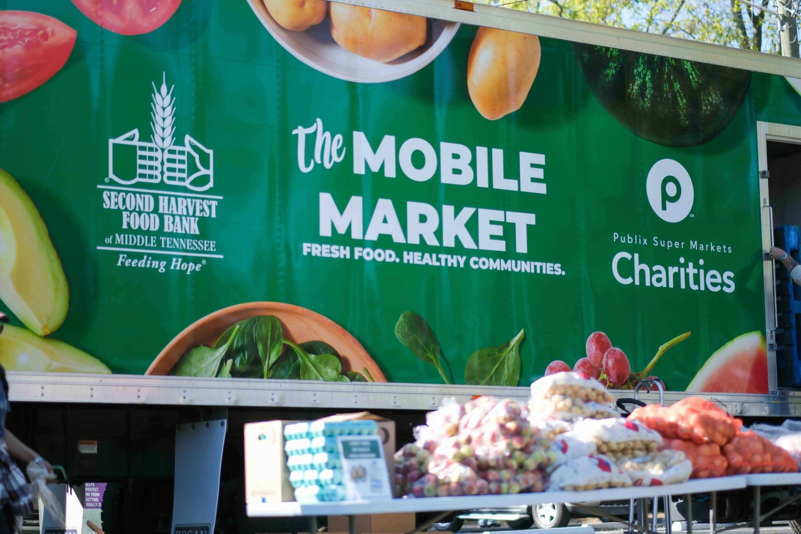 A green truck with The Mobile Market and Second Harvest Food Bank of Middle Tennessee logos is parked, with fresh produce like potatoes, onions, and squash displayed on a table in the foreground.