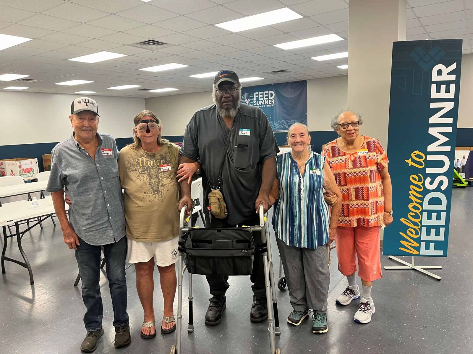 Five adults stand together smiling inside a community center near a Welcome to FEED SUMNER sign. One man uses a walker; the group appears happy and connected, suggesting a supportive event or gathering.