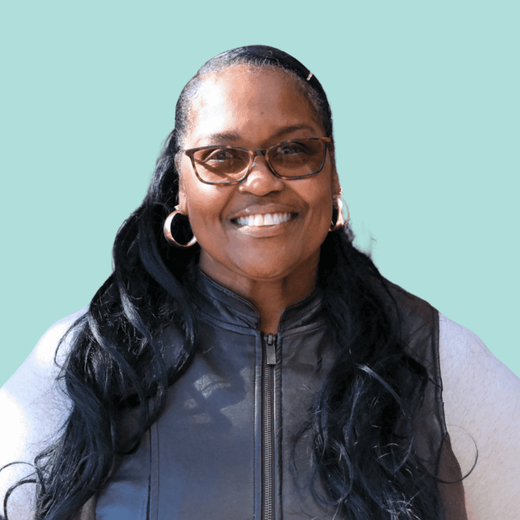 A woman with long black hair, glasses, and hoop earrings smiles while wearing a black vest over a light shirt, standing against a light blue background.