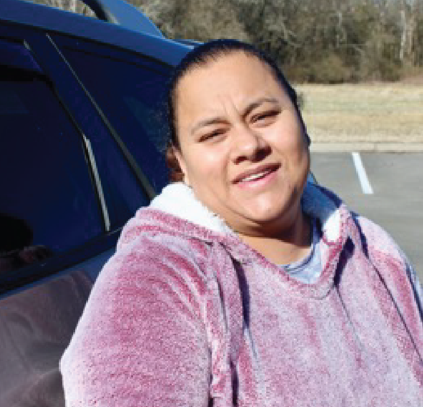 A woman wearing a light purple fleece jacket smiles while standing next to a car in a parking lot on a sunny day, with trees and grass in the background.