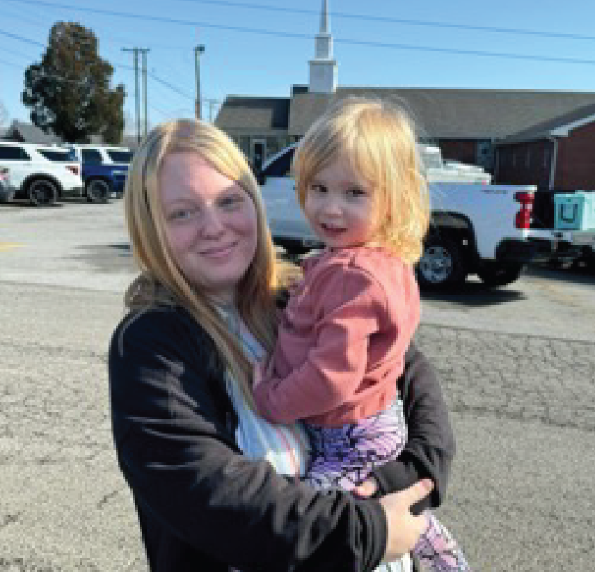 A woman with blonde hair holds a young girl in her arms in a parking lot. Several vehicles and a brick building with a steeple are visible in the background on a sunny day.