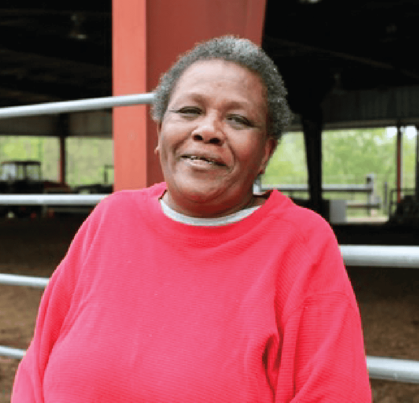 A woman with short curly hair, wearing a bright red sweater, stands and smiles in front of a metal fence with a covered area and greenery in the background.