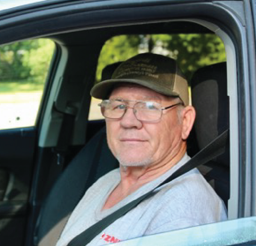 An older man wearing glasses, a gray t-shirt, and a green baseball cap sits in the drivers seat of a car with the window down, looking toward the camera and smiling slightly.