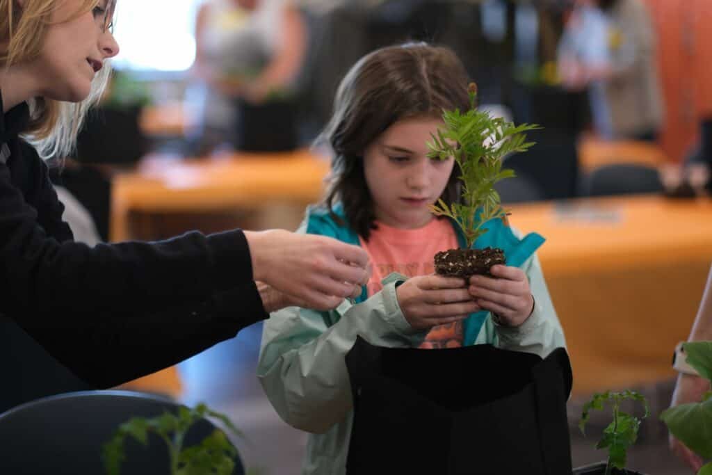 A young girl examines a small plant while an adult assists her. They are indoors at a table with other plants and blurred people in the background, suggesting a group gardening activity or workshop.