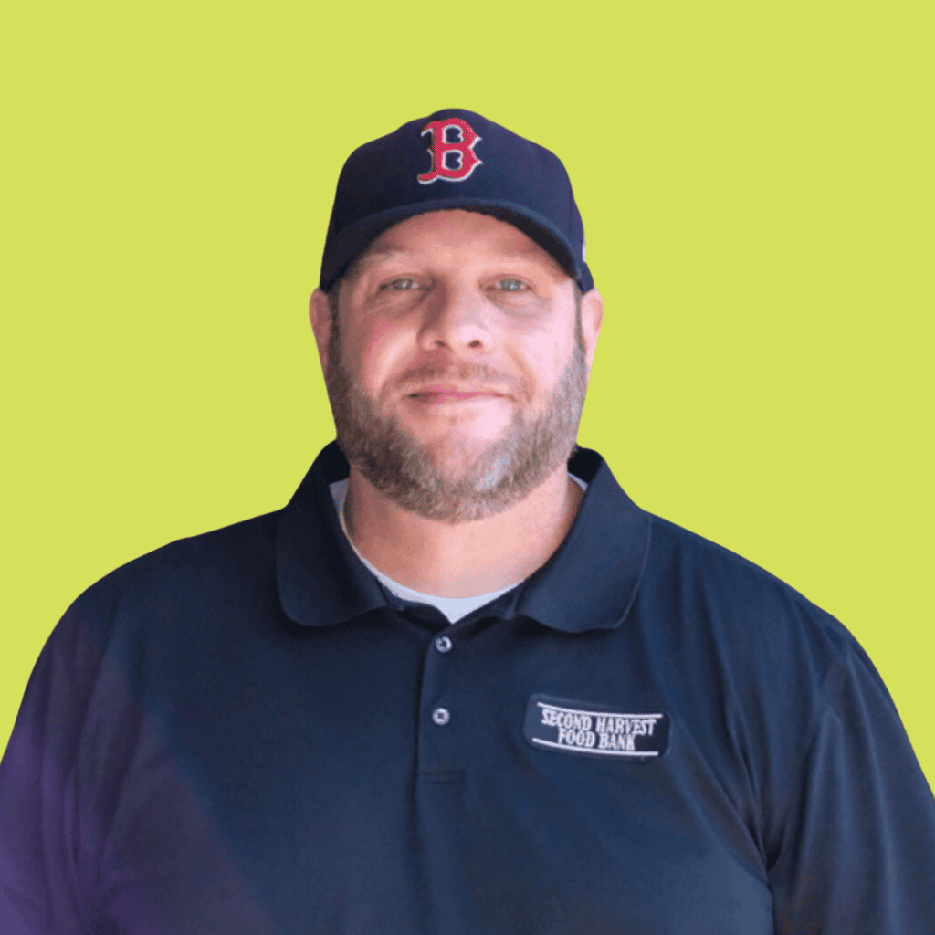 A man with a beard wearing a navy Boston Red Sox baseball cap and a black Second Harvest Food Bank polo shirt stands in front of a light green background.
