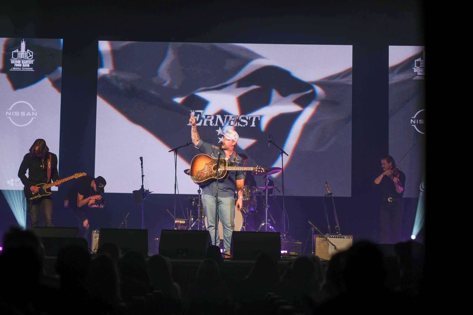 A country musician performs on stage holding a guitar, with a large screen displaying the name ERNEST and a flag background behind him. Band members play instruments under colorful stage lights.