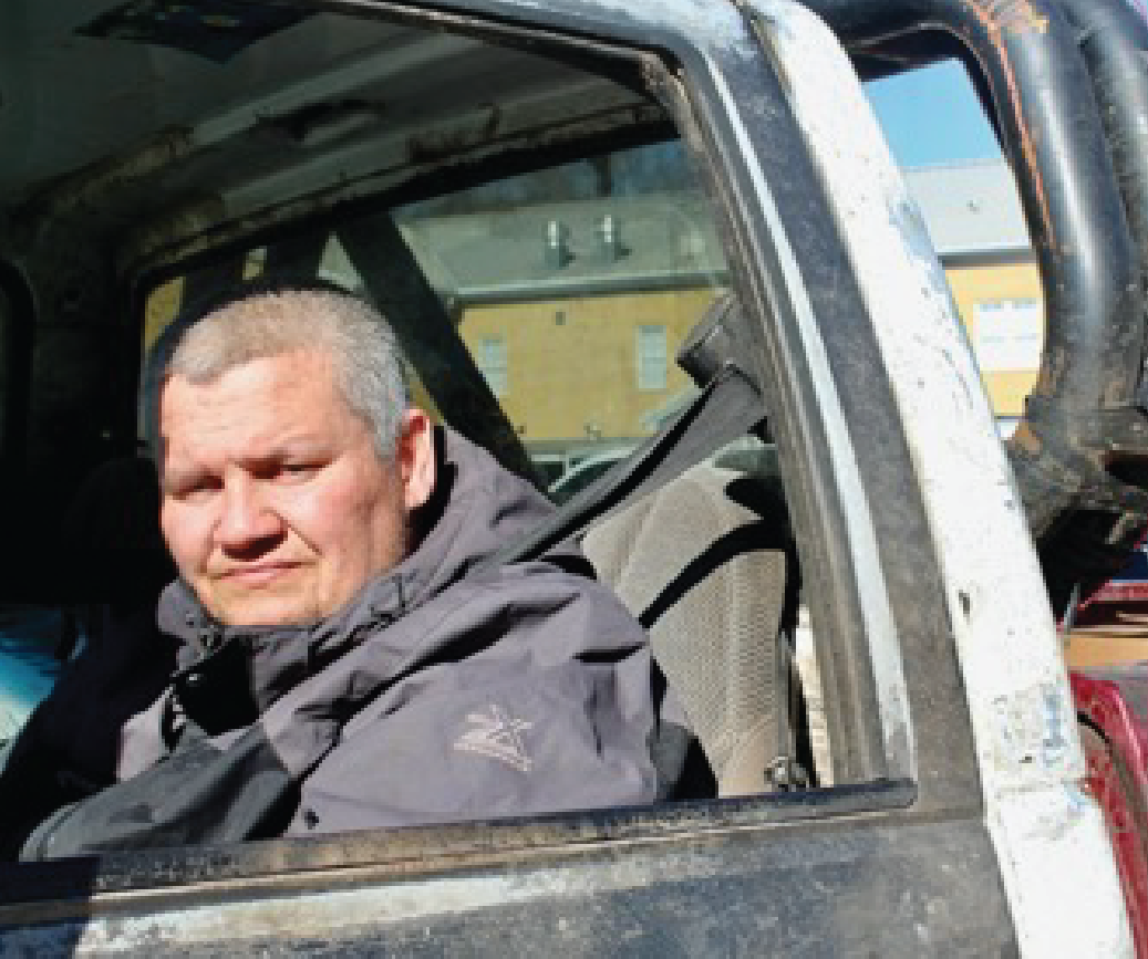 A person with short hair and a dark jacket sits in the back seat of a vehicle, looking out of the open window. Sunlight illuminates their face, and buildings are visible outside.