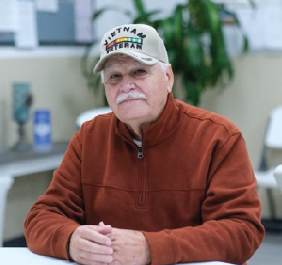 An older man wearing a Vietnam Veteran cap and an orange-brown zip-up jacket sits at a table, clasping his hands. There are blurred office or community room elements in the background.