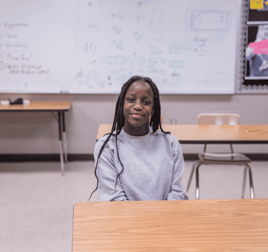 A girl with long braided hair and a gray sweatshirt sits at a desk in a classroom with a whiteboard and empty desks in the background.