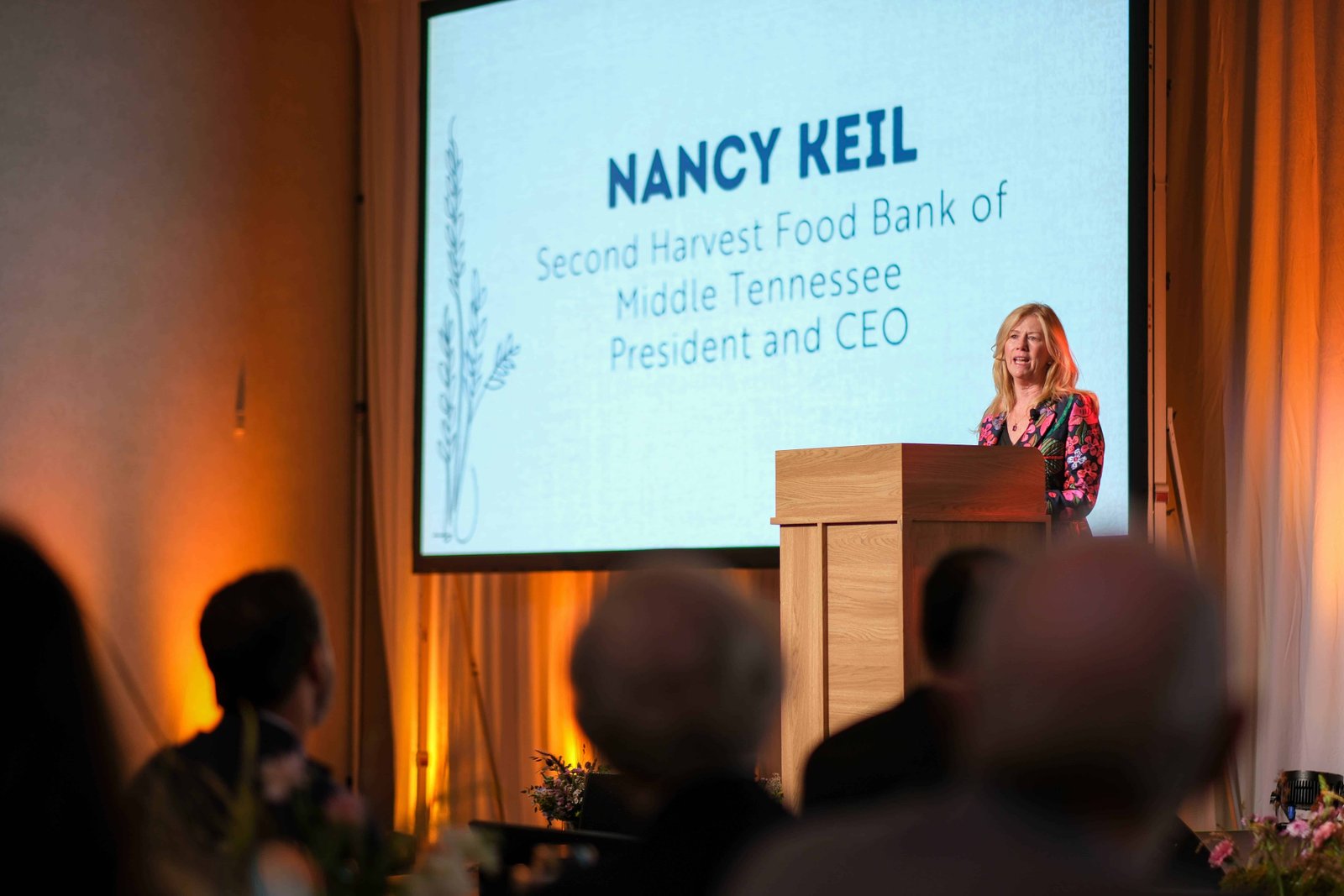 A woman stands at a podium speaking to an audience. Behind her, a large screen displays the text: Nancy Keil, Second Harvest Food Bank of Middle Tennessee, President and CEO.