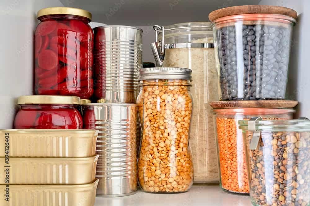 Jars of beans, lentils, grains, and preserves are neatly arranged on a pantry shelf. The dry goods are stacked and organized for easy storage, ready for use or donation to a second harvest.