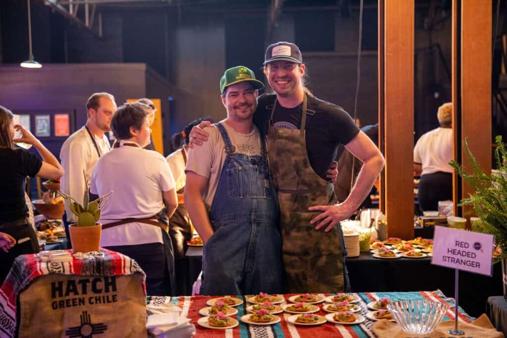 Two smiling men in aprons stand side by side behind a table with prepared food plates at an indoor event. The table is covered with a colorful cloth and labeled “Red Headed Stranger.” Other people and food vendors are in the background.