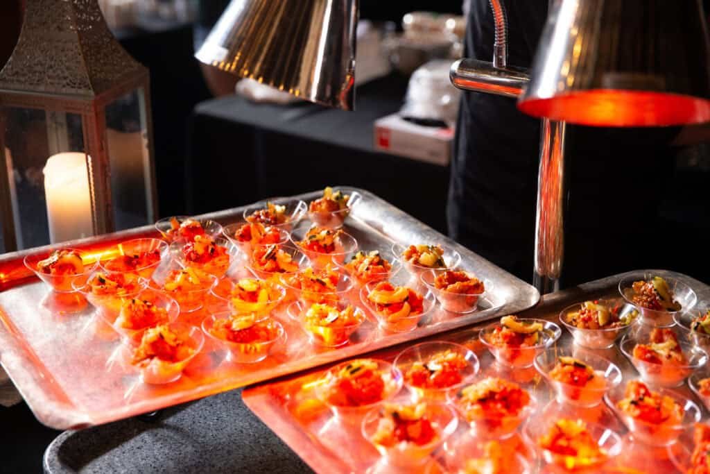 Two trays of small appetizer cups are arranged under warming lamps, each cup filled with colorful bite-sized food portions. The trays are set on a counter in a dimly lit event or catering setting.