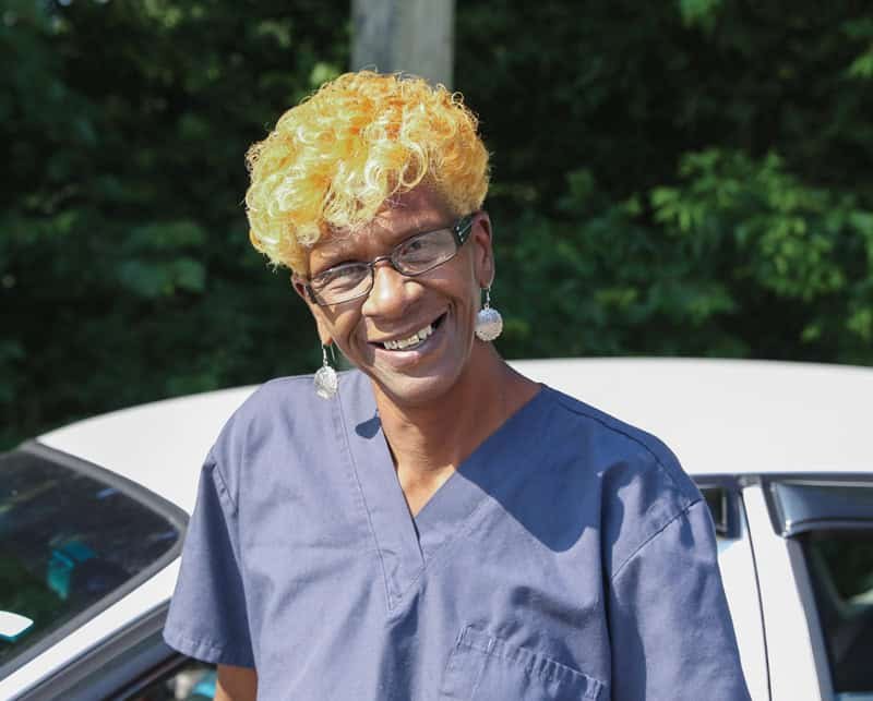 A person with short curly blonde hair, glasses, and dangling earrings smiles while wearing blue scrubs, standing outdoors in front of a white car with greenery in the background.