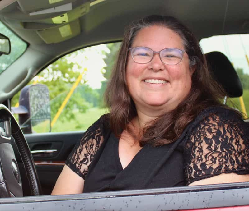 A woman with long brown hair and glasses sits in the driver’s seat of a car, smiling. She wears a black top with lace sleeves. The car window is open, and greenery is visible outside.
