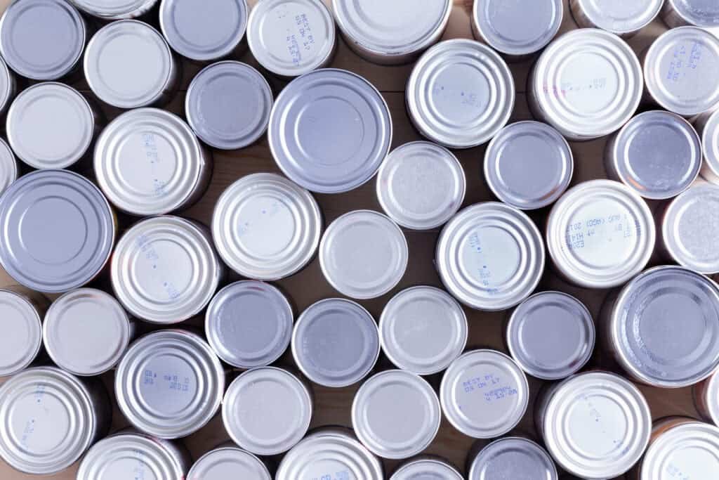 A top-down view of many silver and white tin cans arranged closely together, showing their lids and stamped dates or codes. The variety of sizes highlights how healthy food can be simple and affordable when choosing canned options.