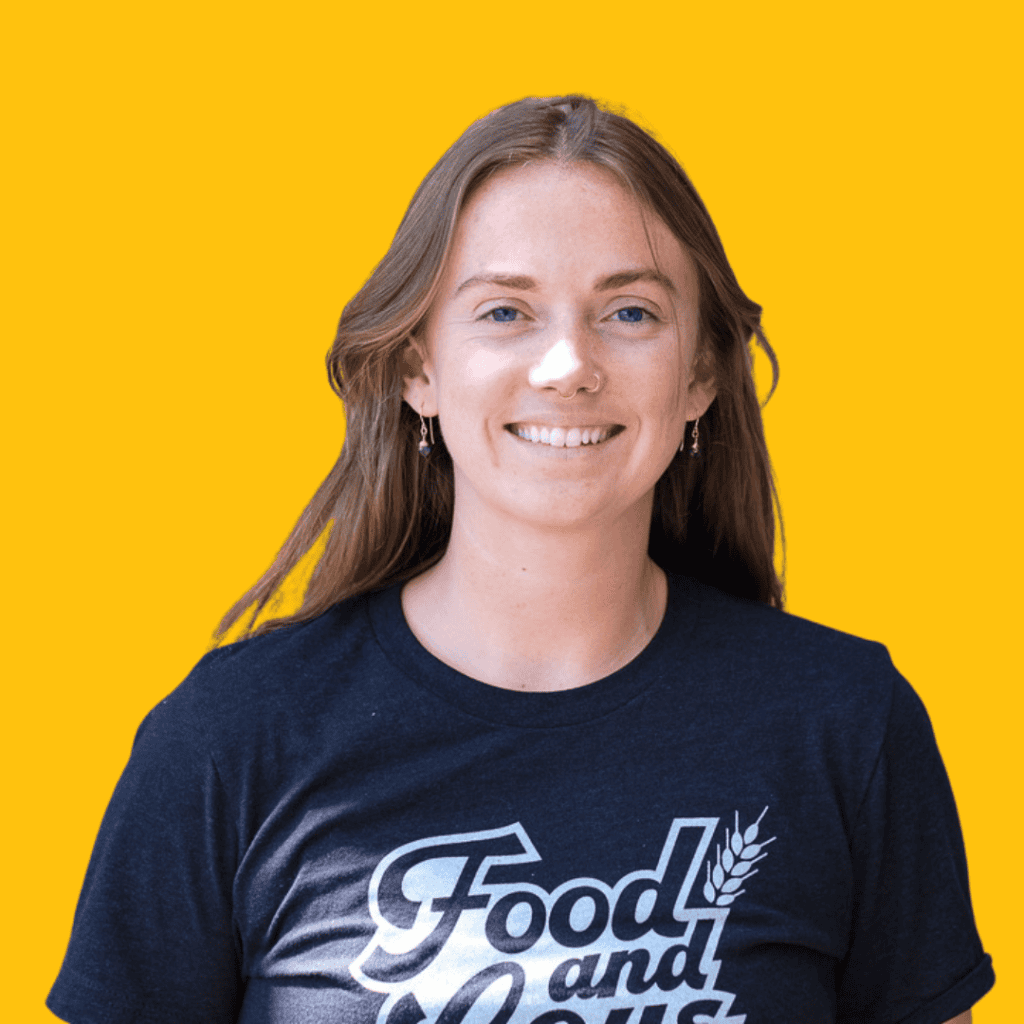 A young woman with long brown hair smiles at the camera. She is wearing a navy blue Food and t-shirt and standing in front of a solid bright yellow background.