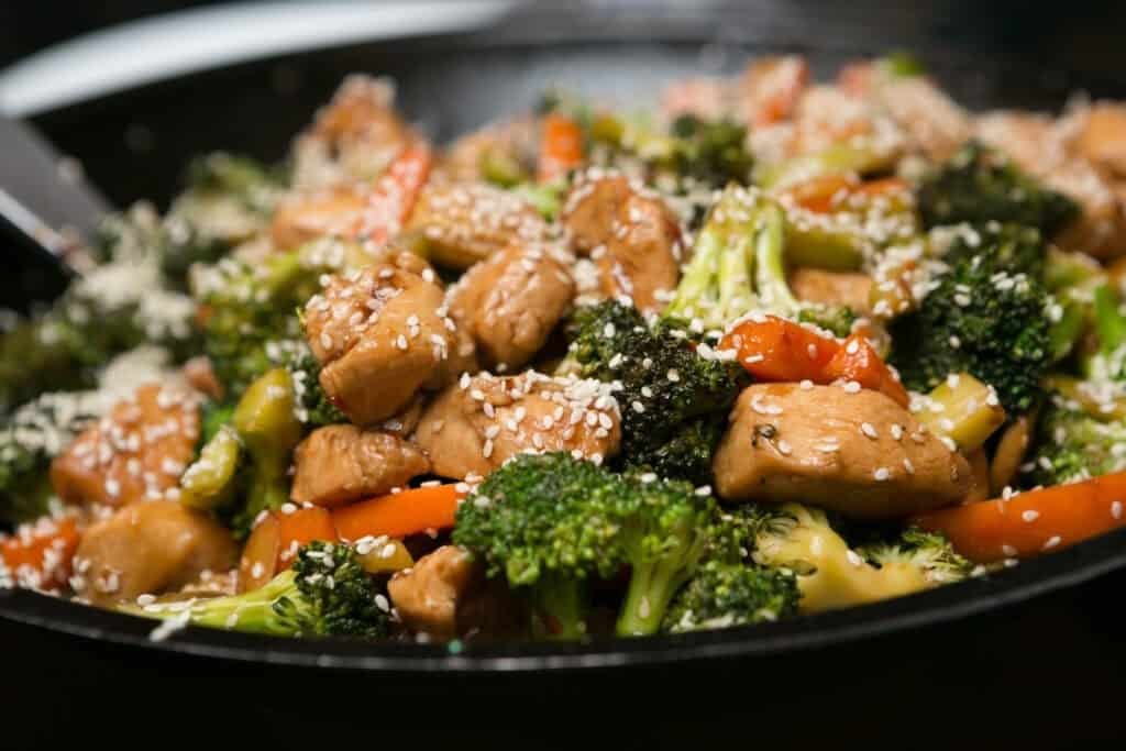 A close-up of a stir-fry dish featuring chicken, broccoli, red bell peppers, and sesame seeds in a black pan. This fresh and colorful meal is a great example of healthy and affordable meals.