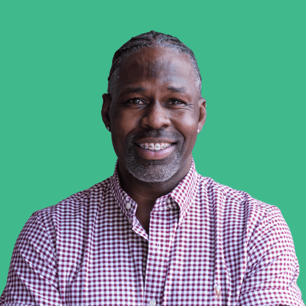 A man with short gray hair and a trimmed beard smiles at the camera. He is wearing a red and white checkered shirt and stands in front of a solid teal background.
