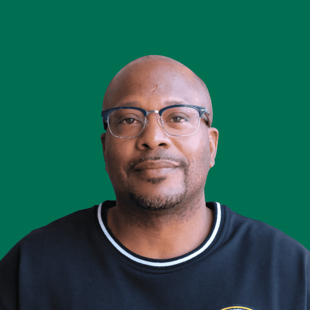 A man with glasses and a short beard wearing a black shirt stands in front of a solid green background, looking at the camera with a neutral expression.