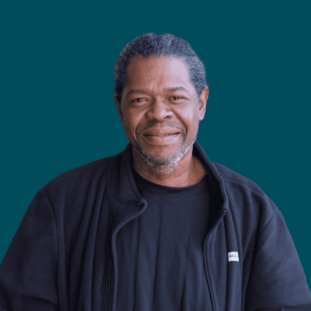 A middle-aged man with short curly hair and a beard, wearing a black jacket and a black shirt, smiles gently while standing against a solid teal background.