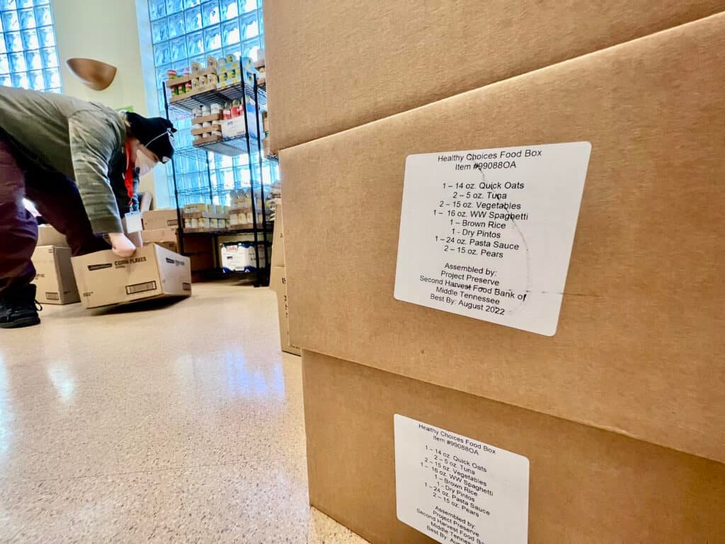 A person in a mask moves boxes in a food bank. In the foreground, “Healthy Choices Food Box” labels highlight items for eating healthy on a budget for blood pressure control. Shelves of groceries are visible in the background.
