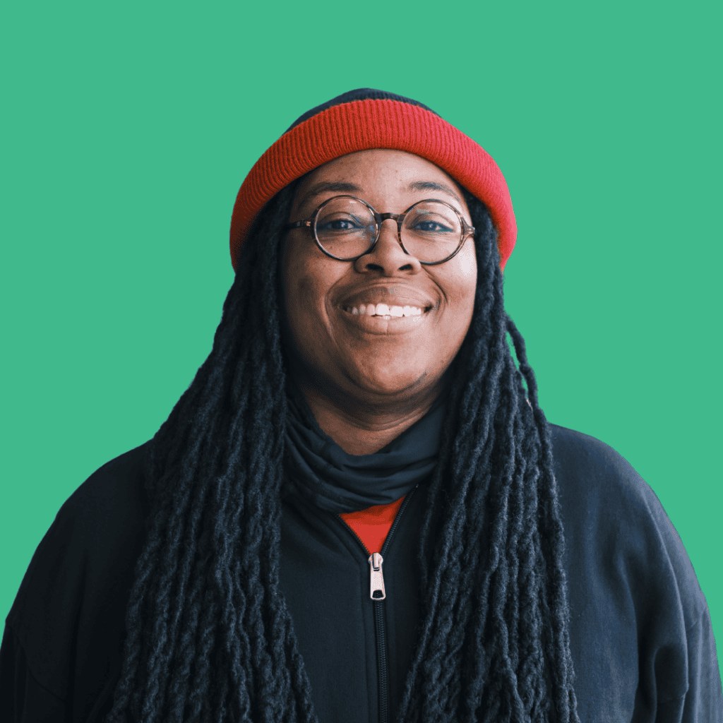 A person with long black locs, wearing glasses, a red beanie, and a black jacket, smiling in front of a solid teal background.