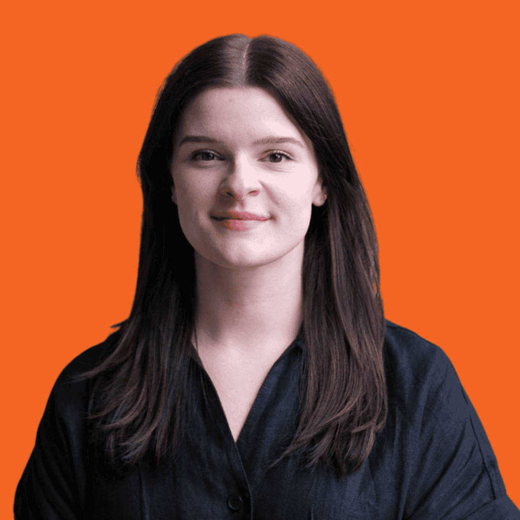 A young woman with straight brown hair, wearing a black button-up shirt, smiles slightly while standing in front of a plain bright orange background.