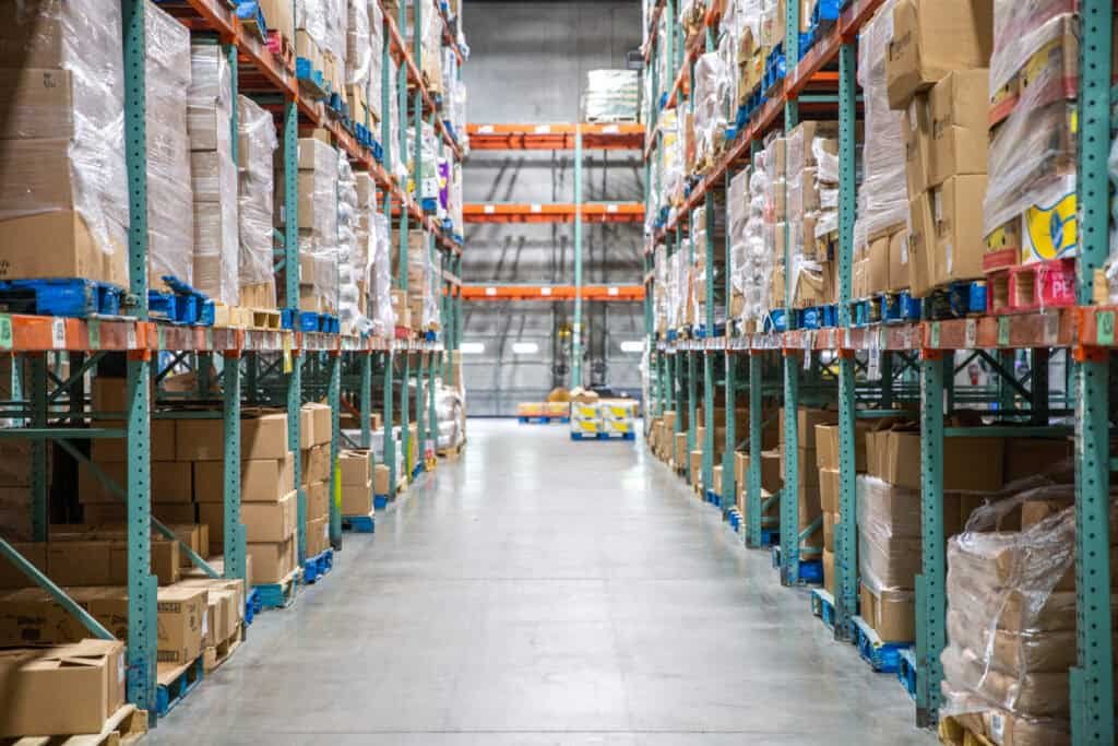 A warehouse aisle lined with tall shelves stacked with boxes and pallets, extending into the distance under bright overhead lighting. The floor is clear and clean.