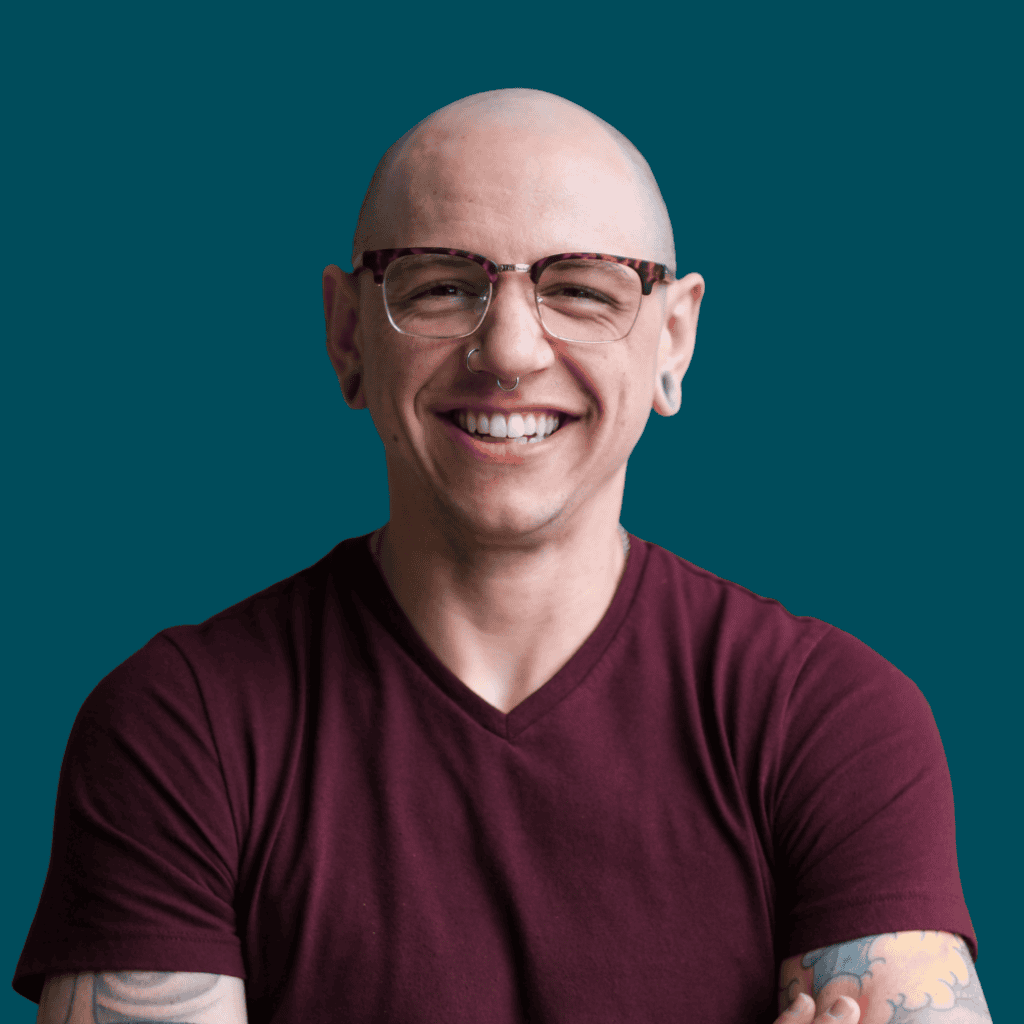 A smiling person with glasses, a nose ring, and visible tattoos on their forearm, wearing a maroon t-shirt, standing against a solid teal background.