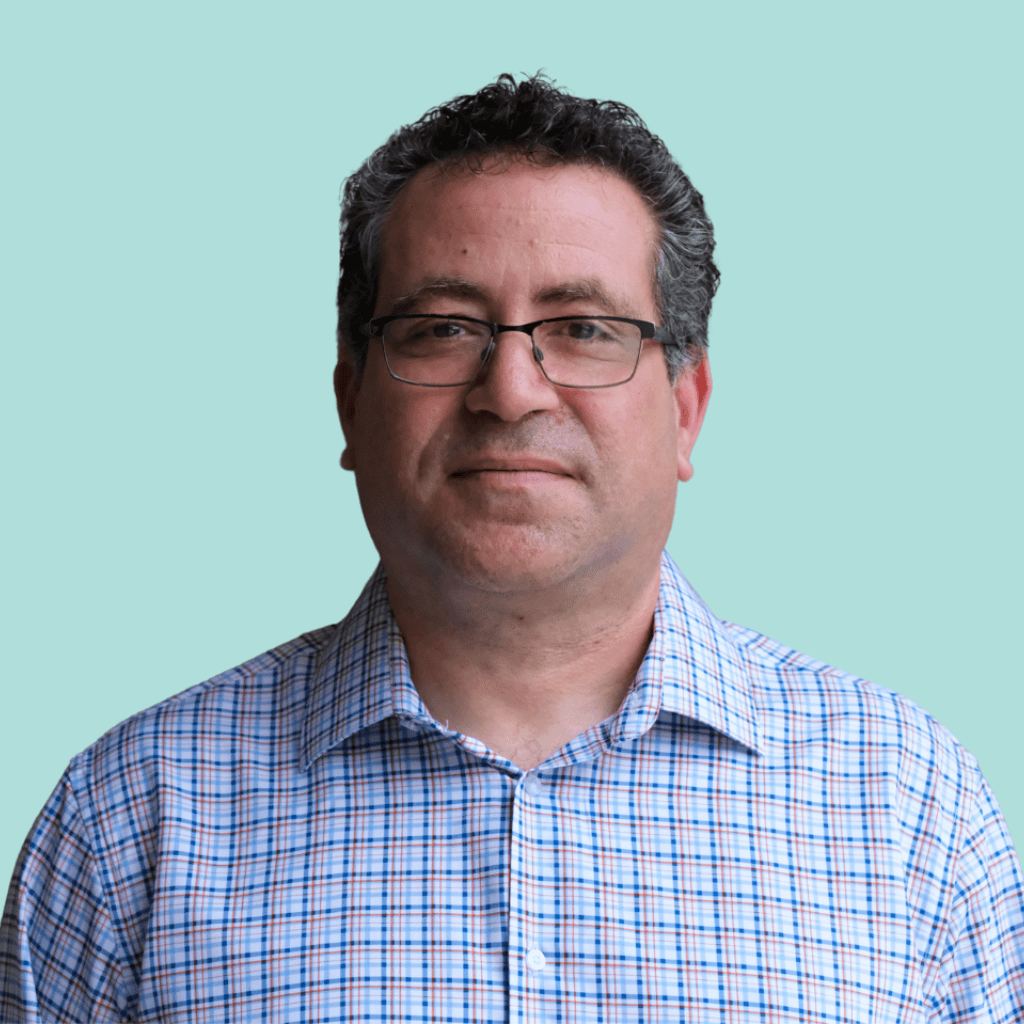 A man with short curly hair and glasses is wearing a light blue and pink checkered shirt, standing against a plain light blue background. He is looking straight at the camera with a neutral expression.