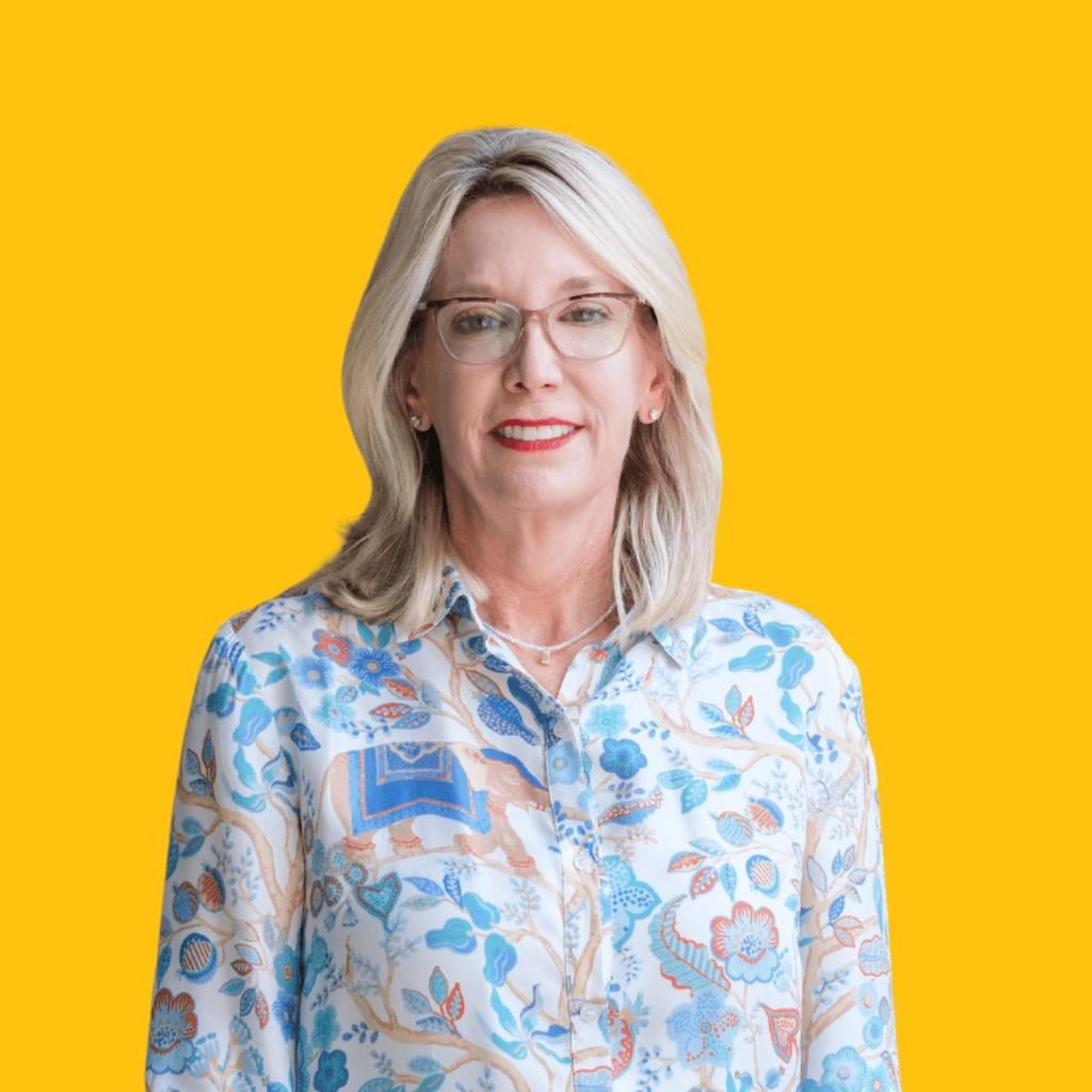 A smiling woman with blonde hair and glasses wearing a colorful floral blouse stands in front of a bright yellow background.