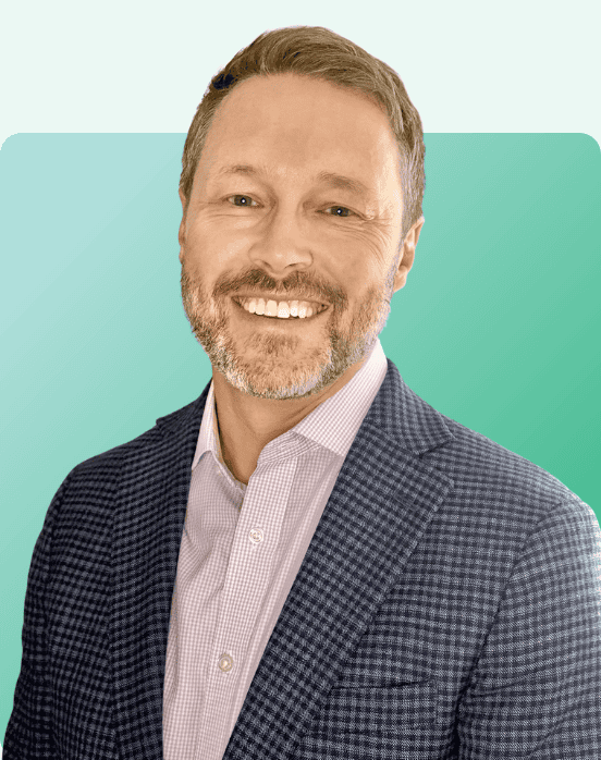 A smiling man with short light brown hair and a beard, wearing a checked navy blazer over a light purple shirt, poses in front of a teal gradient background.
