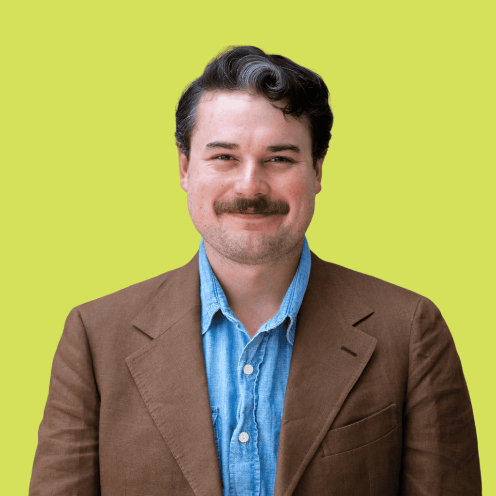 A man with dark hair and a mustache, wearing a brown blazer over a light blue shirt, stands smiling in front of a plain yellow background.